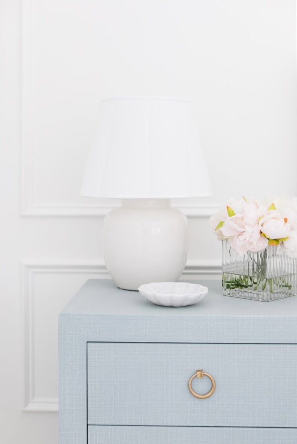 A white table lamp, a glass vase with pink flowers, and a small white dish sit on a light blue drawer with a gold ring handle, offering charming nightstand decorating inspiration against a crisp white wall.