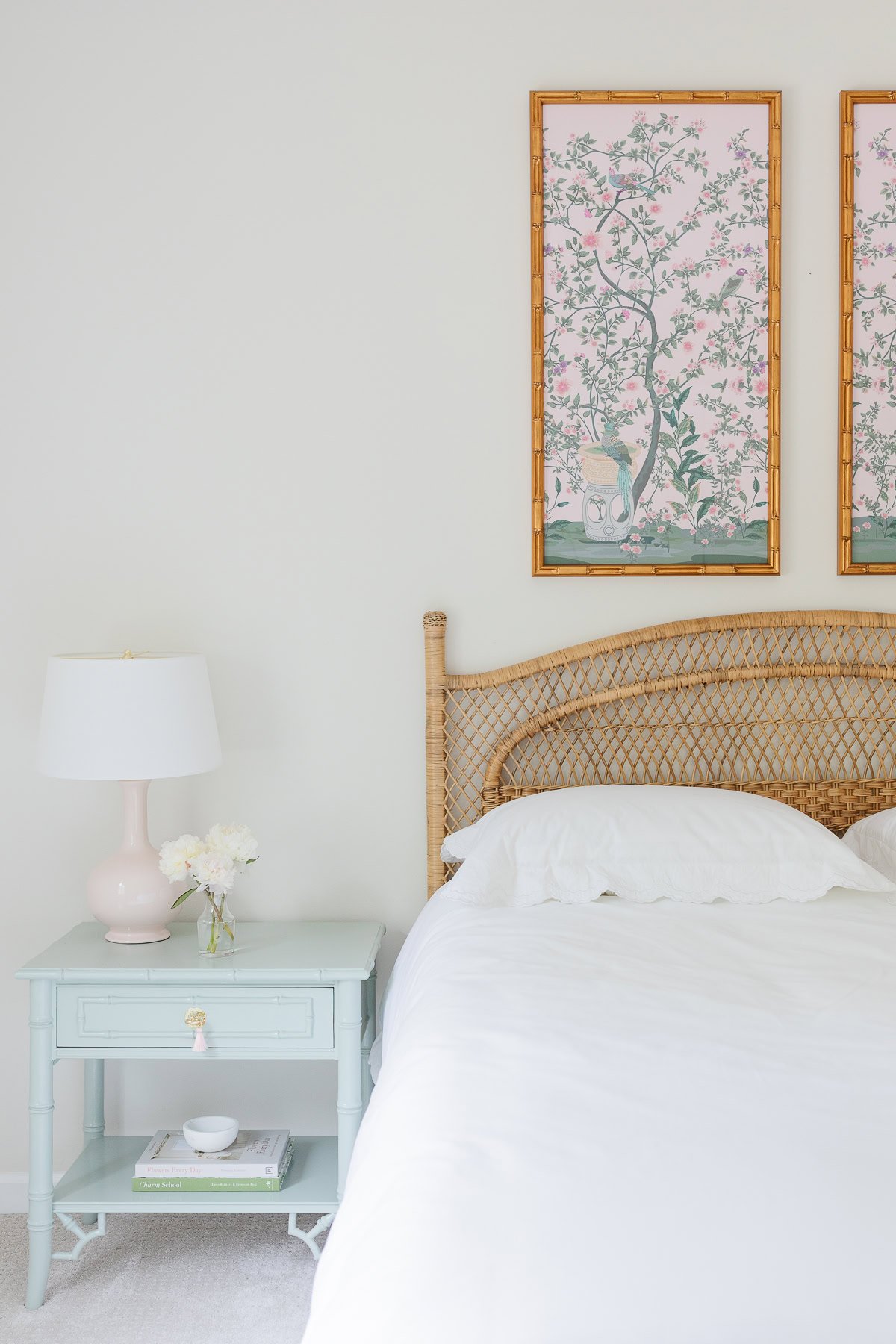 A neatly made bed with a wicker headboard, a light blue nightstand decorated with books, flowers, and a white lamp—perfect for nightstand decorating—and two framed botanical prints displayed above.