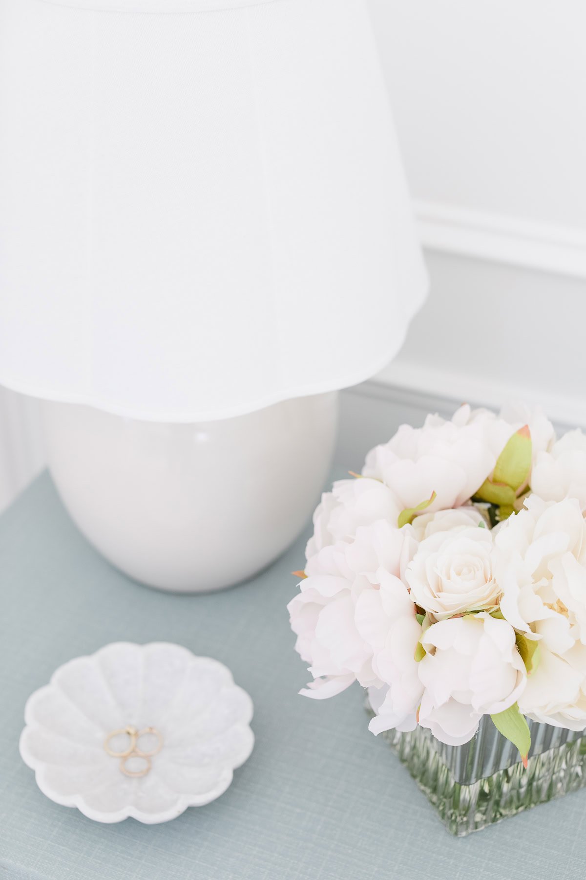 A white lamp, a glass vase with white flowers, and a small dish holding two rings are displayed on a light blue textured surface—perfect inspiration for nightstand decorating.