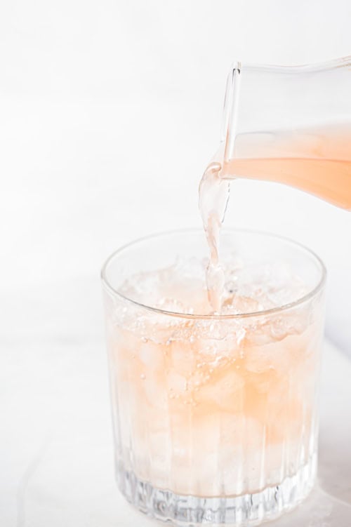Grapefruit soda being poured into a paloma cocktail glass filled with ice cubes on a white background.