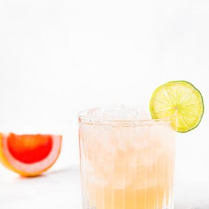 A glass of pink paloma cocktail with a lime slice on the rim, ice cubes, and a halved grapefruit in the background on a light surface.