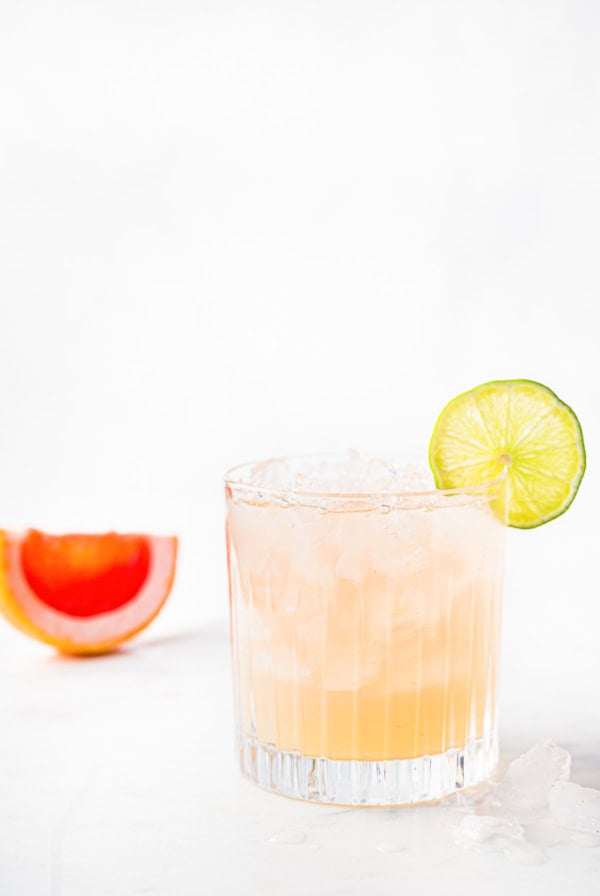 A glass of pink paloma cocktail with a lime slice on the rim, ice cubes, and a halved grapefruit in the background on a light surface.