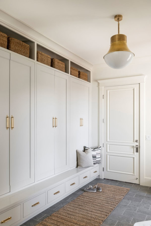 A white mudroom with built-in cabinets, wicker baskets on top, a bench with pillows, and a rug on the floor. The walls are painted in Benjamin Moore Revere Pewter. A brass light fixture hangs from the ceiling.