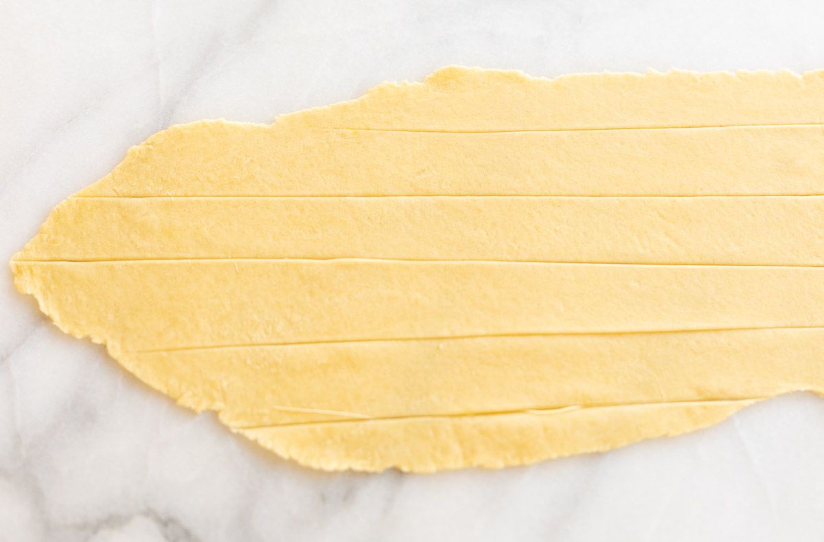 A sheet of homemade pasta dough laid out on a marble surface, cut into strips for Pappardelle