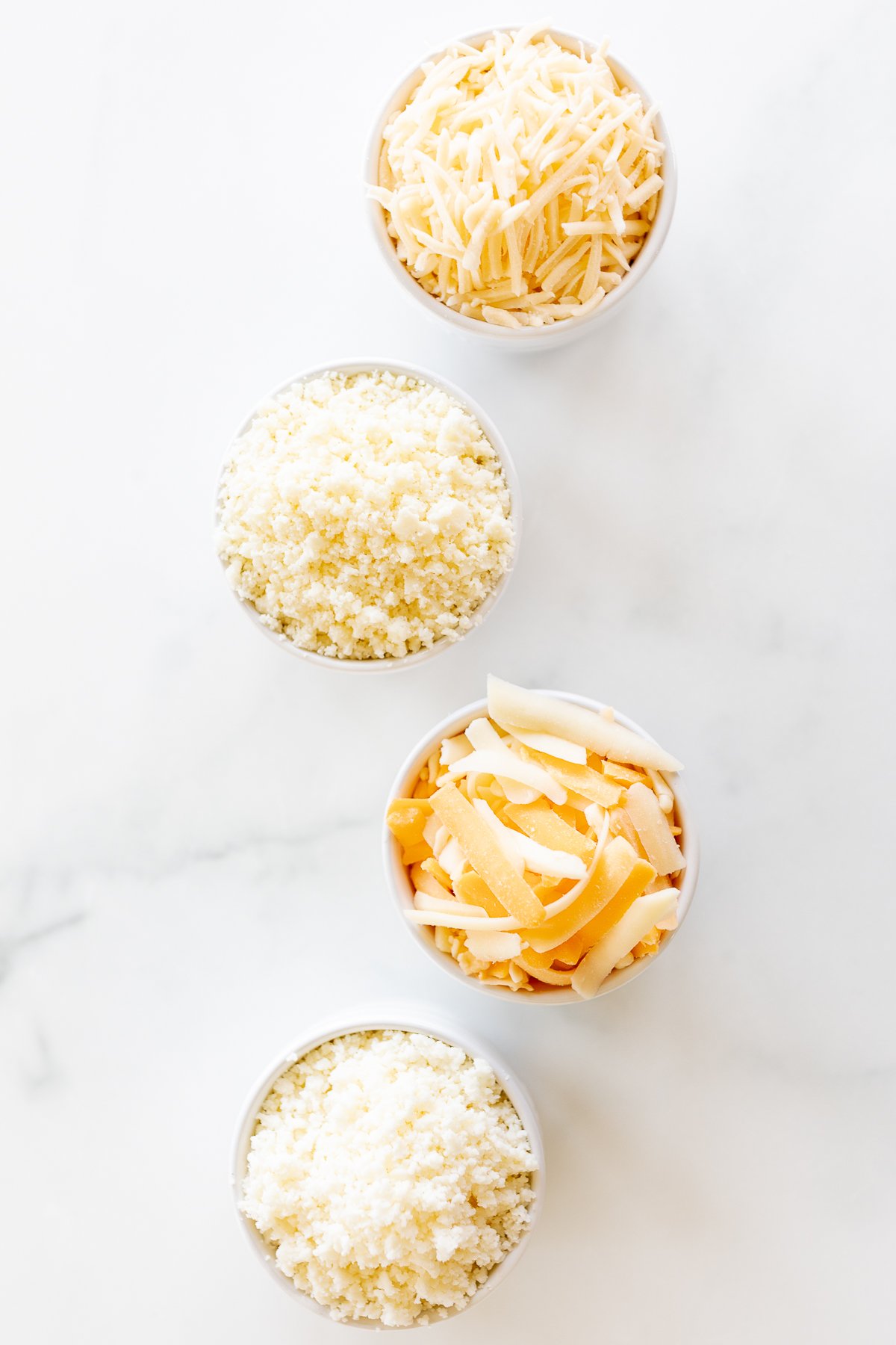 Four bowls of Mexican cheese in white bowls on a marble countertop.