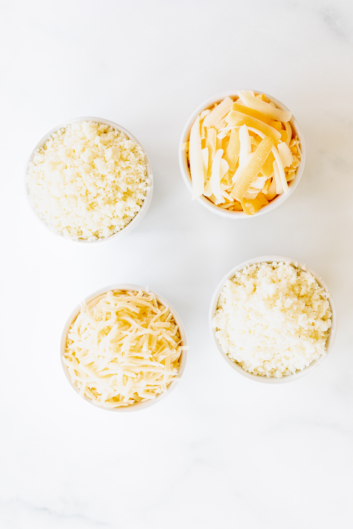 Four bowls of Mexican cheese varieties in white bowls on a marble surface.