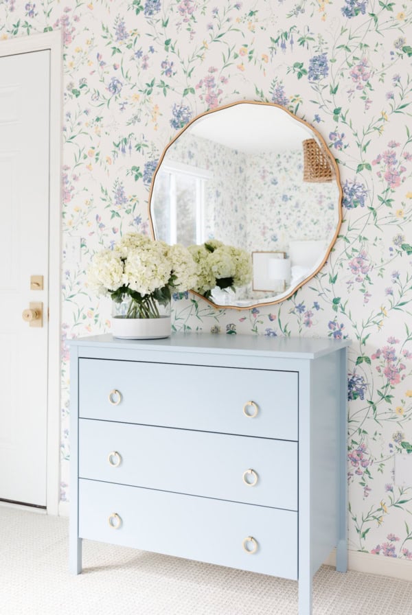 A light blue dresser in coastal blues with three drawers stands against a floral wallpapered wall, topped with white hydrangea flowers in a vase and a large round mirror above.