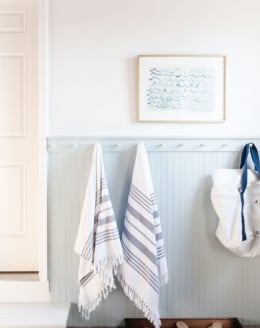 A mudroom area with beadboard painted in a coastal blue color, towels and a beach bag hanging on hooks