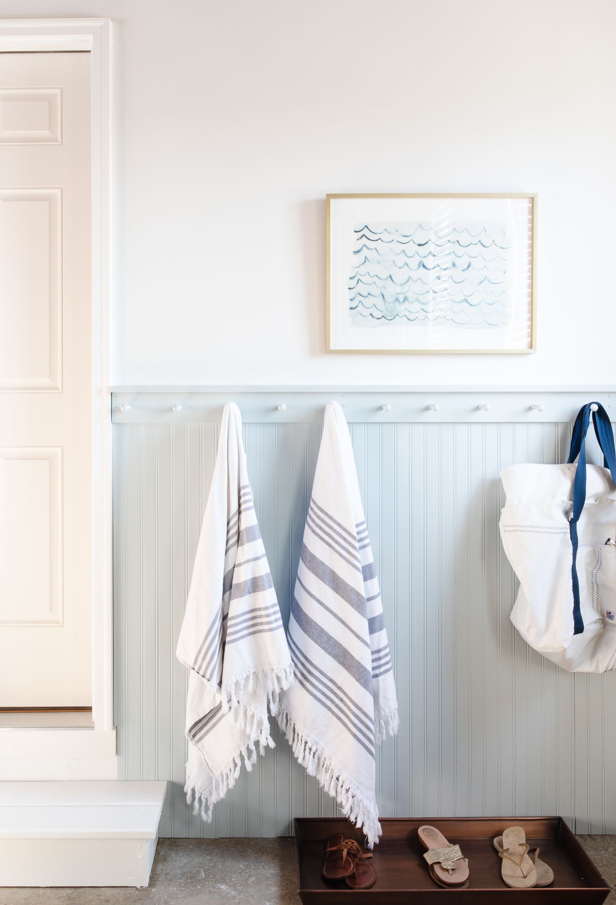 A mudroom area with beadboard painted in a coastal blue color, towels and a beach bag hanging on hooks