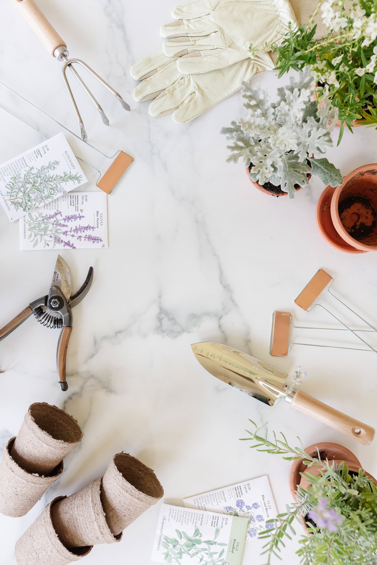 Gardening tools, gloves, plant pots, seed packets, and plant markers arranged on a white marble surface with potted plants—perfect essentials to organize on potting benches.