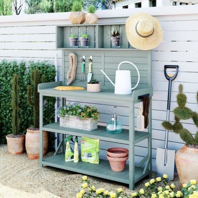Green outdoor potting bench against a white fence, holding gardening tools, watering can, potted plants, supplies, and a straw hat. Potting benches and a hose hang nearby for easy access.