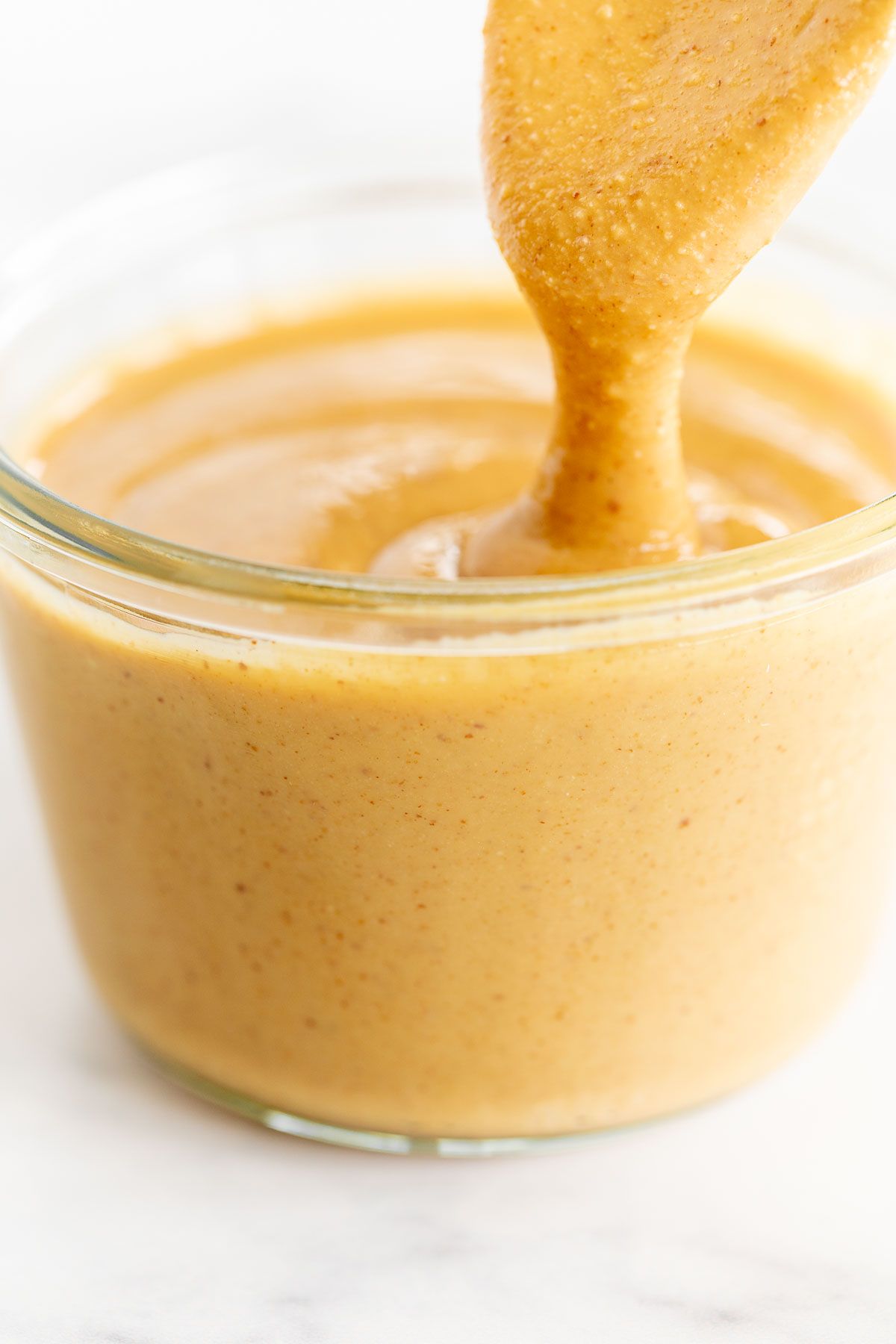 A clear glass jar full of a homemade peanut butter recipe, spoon pulling some out at the top.