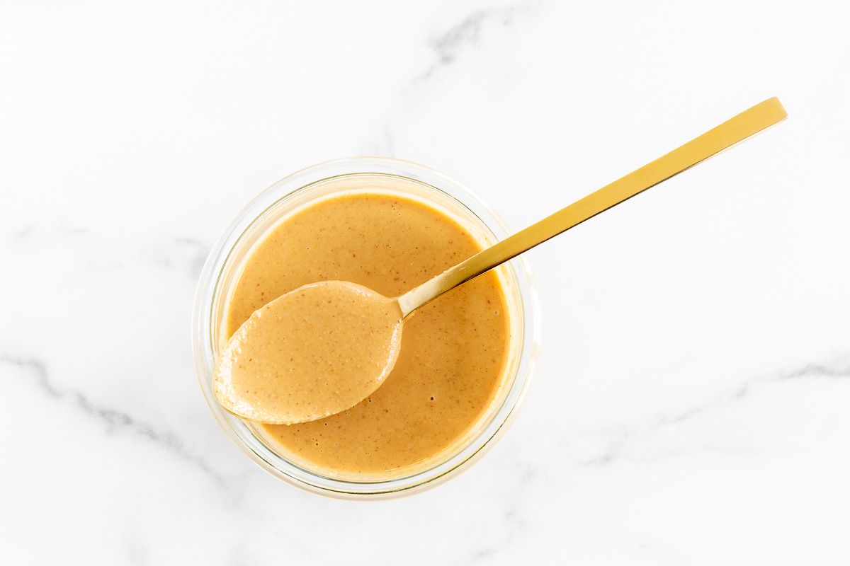 A small glass jar of homemade peanut butter on a marble surface, gold spoon across top.
