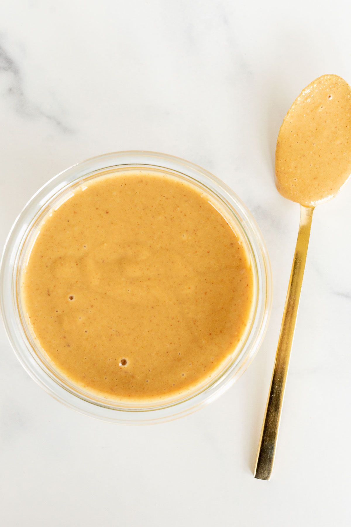 A small glass jar of homemade peanut butter on a marble surface, gold spoon to the side.