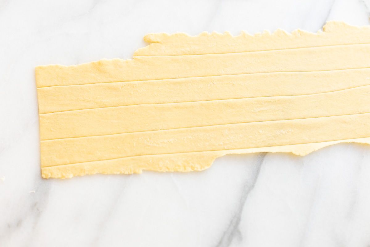 A sheet of homemade pasta dough laid out on a marble surface, cut into strips for Pappardelle