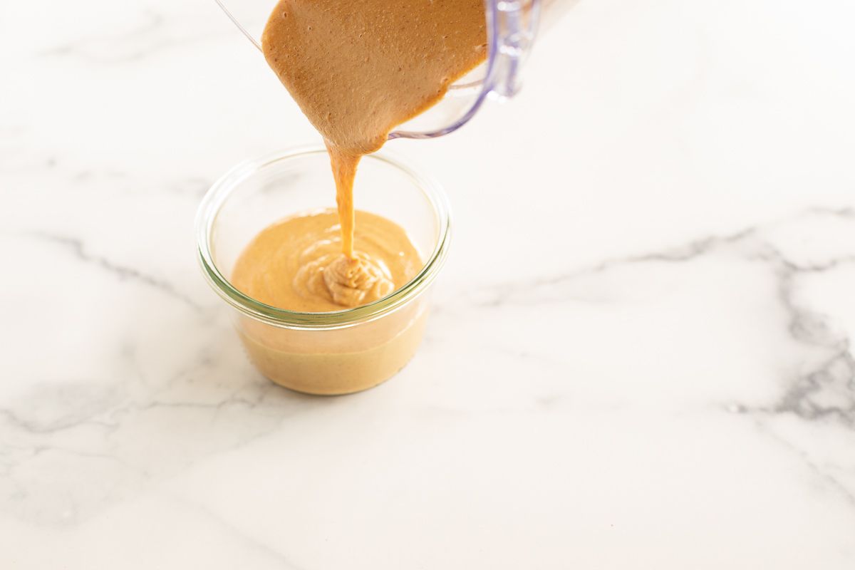Homemade peanut butter pouring into a glass jar from a food processor.