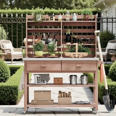A wooden potting bench with drawers, shelves, and gardening tools is set up outdoors, holding potted plants and supplies. Other potting benches, patio furniture, and trimmed hedges complete the inviting garden scene.