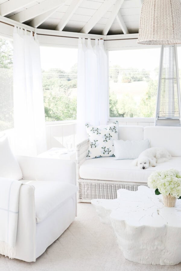 A bright sunroom built with screen porch kits, featuring white furniture, a white dog sleeping on a sofa, and a floral-patterned throw pillow.