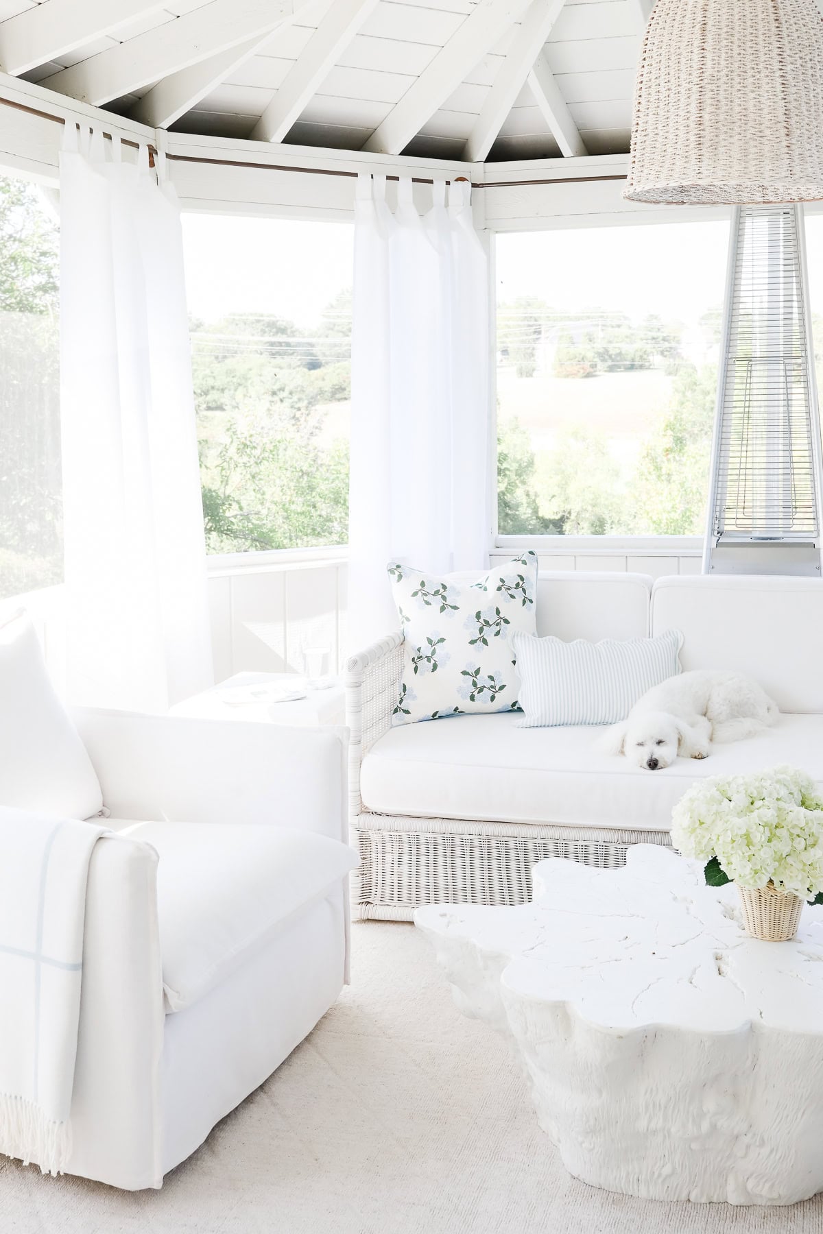 A white sunroom built from screen porch kits, furnished with a sofa, armchair, and coffee table, featuring a small white dog sleeping on the sofa.