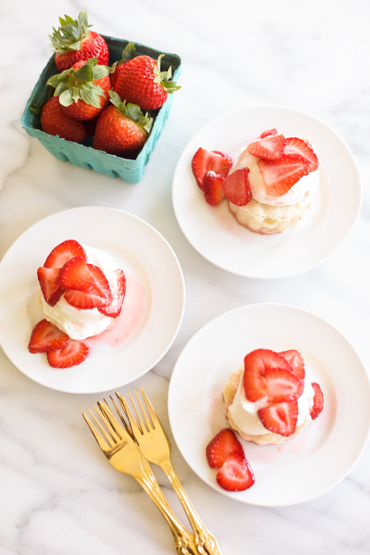 Small plates of strawberry shortcake served with whipped cream and sliced strawberries, set next to a basket of fresh strawberries and three golden forks on a marble surface. This delightful setup is perfect for those seeking exquisite strawberry recipes.