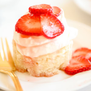 A slice of shortcake topped with whipped cream and fresh strawberry slices, placed on a white plate next to a golden fork, showcases the best in traditional strawberry recipes.