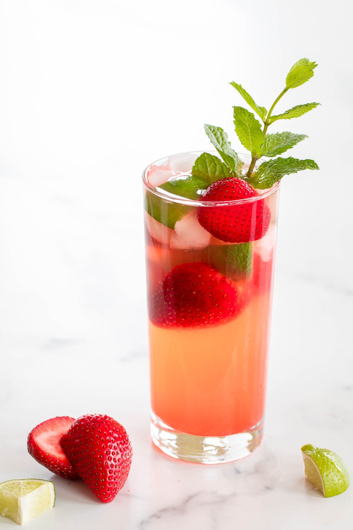 A tall glass filled with a pink drink garnished with whole strawberries, ice cubes, a slice of lime, and a sprig of mint. Showcasing one of the most refreshing strawberry recipes, this delightful beverage is complemented by two strawberry halves and a lime wedge on the white surface next to the glass.