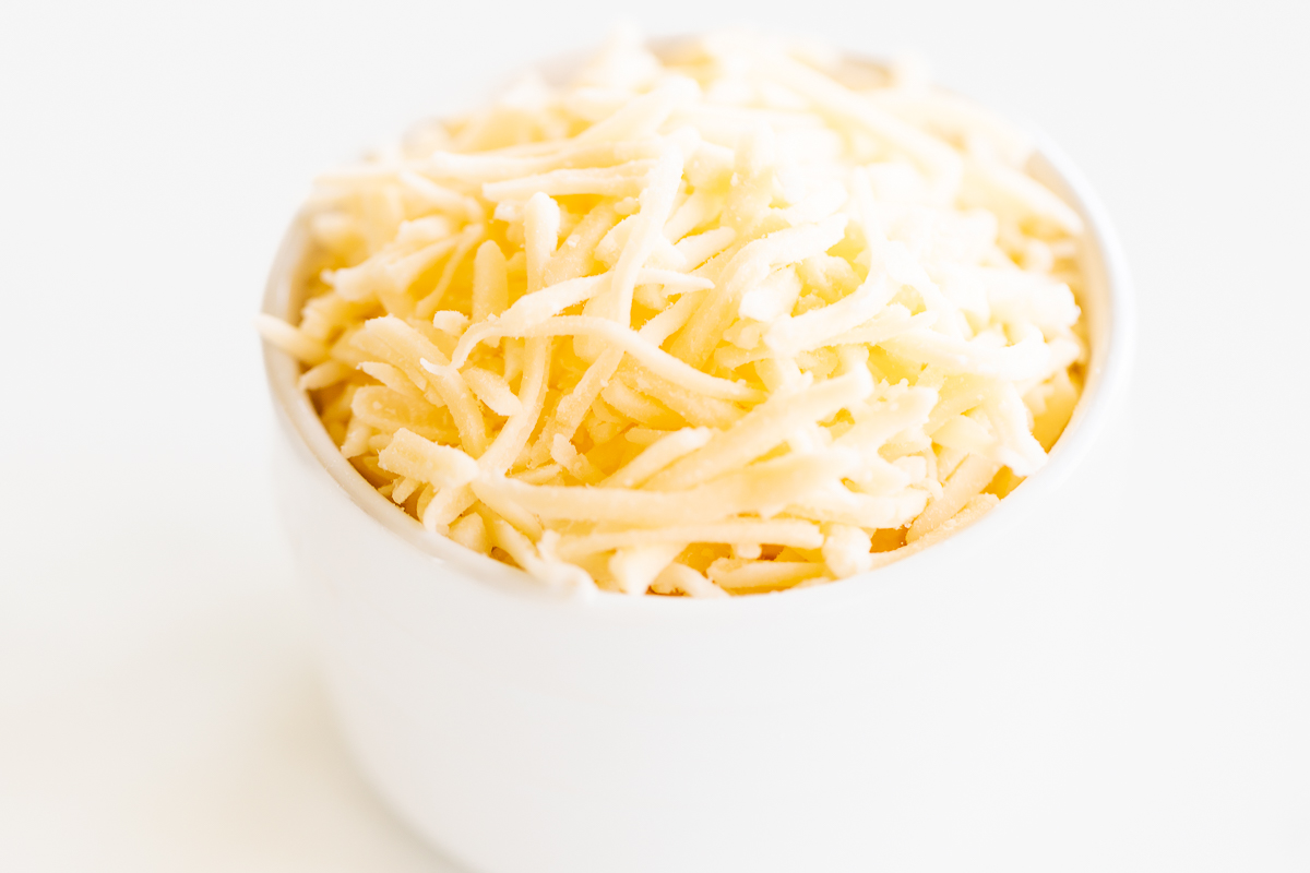 Shredded Mexican cheese in a white cup on a white surface.