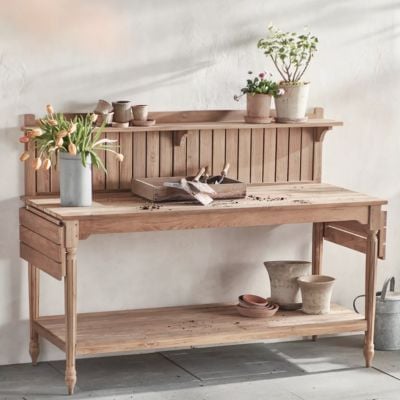 Wood potting bench with gardening tools, potted plants, and flowers arranged on top and below, showcasing the practicality of potting benches against a light-colored wall.