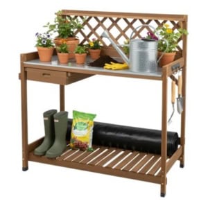 A wooden potting bench holds potted plants, gardening tools, a watering can, gloves, rubber boots, a bag of potting mix, and garden fabric—perfect for anyone who loves potting benches and organized garden spaces.