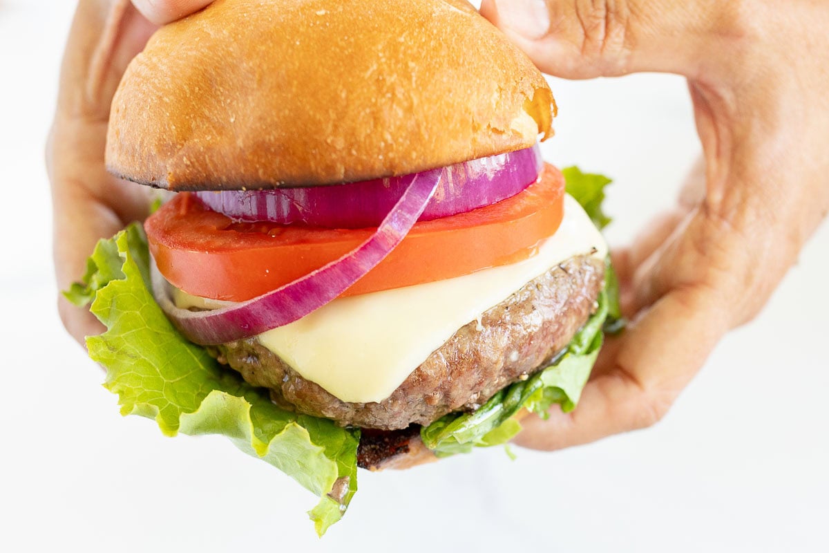 A person holding a hamburger with lettuce, a beef patty, melted cheese, tomato slices, red onion rings, and a bun—perfectly paired with your favorite burger sides.