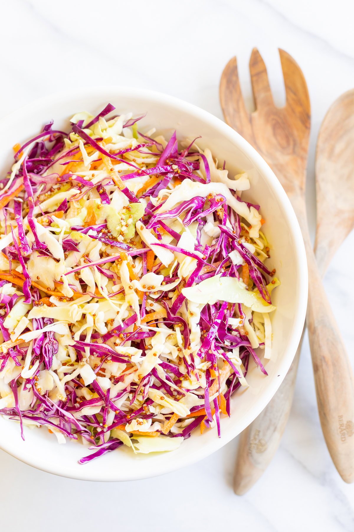 A bowl of colorful coleslaw with shredded cabbage and carrots, perfect among burger sides, accompanied by wooden salad serving utensils on a white marble surface.