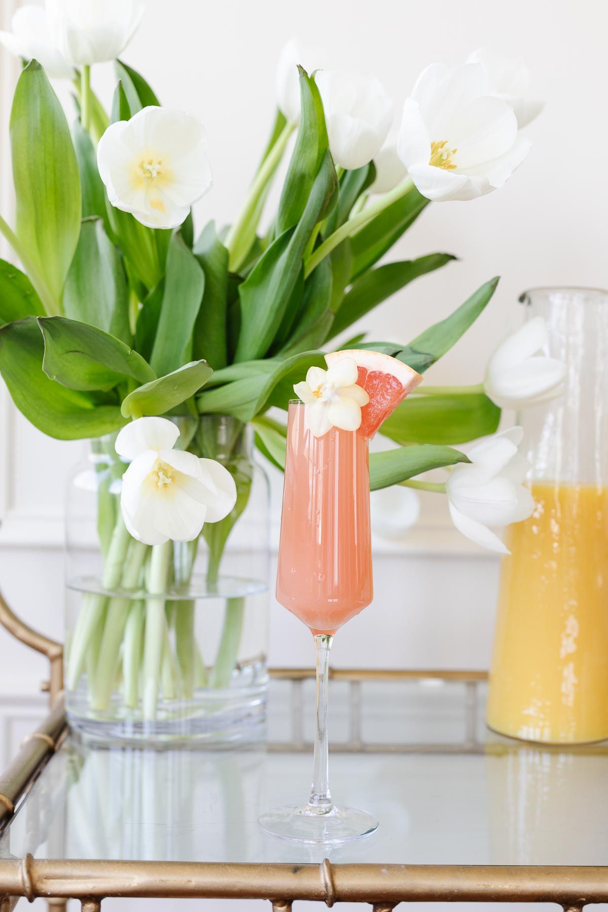 A grapefruit paloma on a gold bar cart, white tulips and fresh juices in the background
