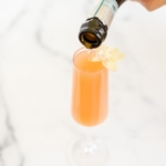 A grapefruit paloma on a marble surface, bottle of champagne to the side