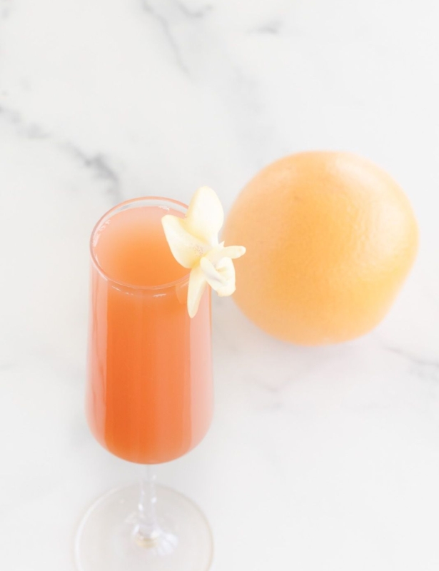 A grapefruit mimosa on a marble surface, garnished with a flower and a whole grapefruit in the background