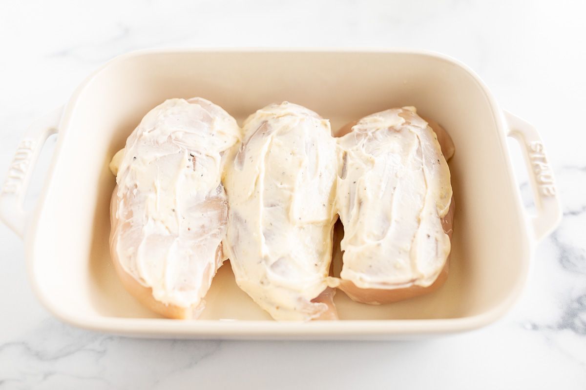 Three chicken breasts covered in mayonnaise in a white baking dish.