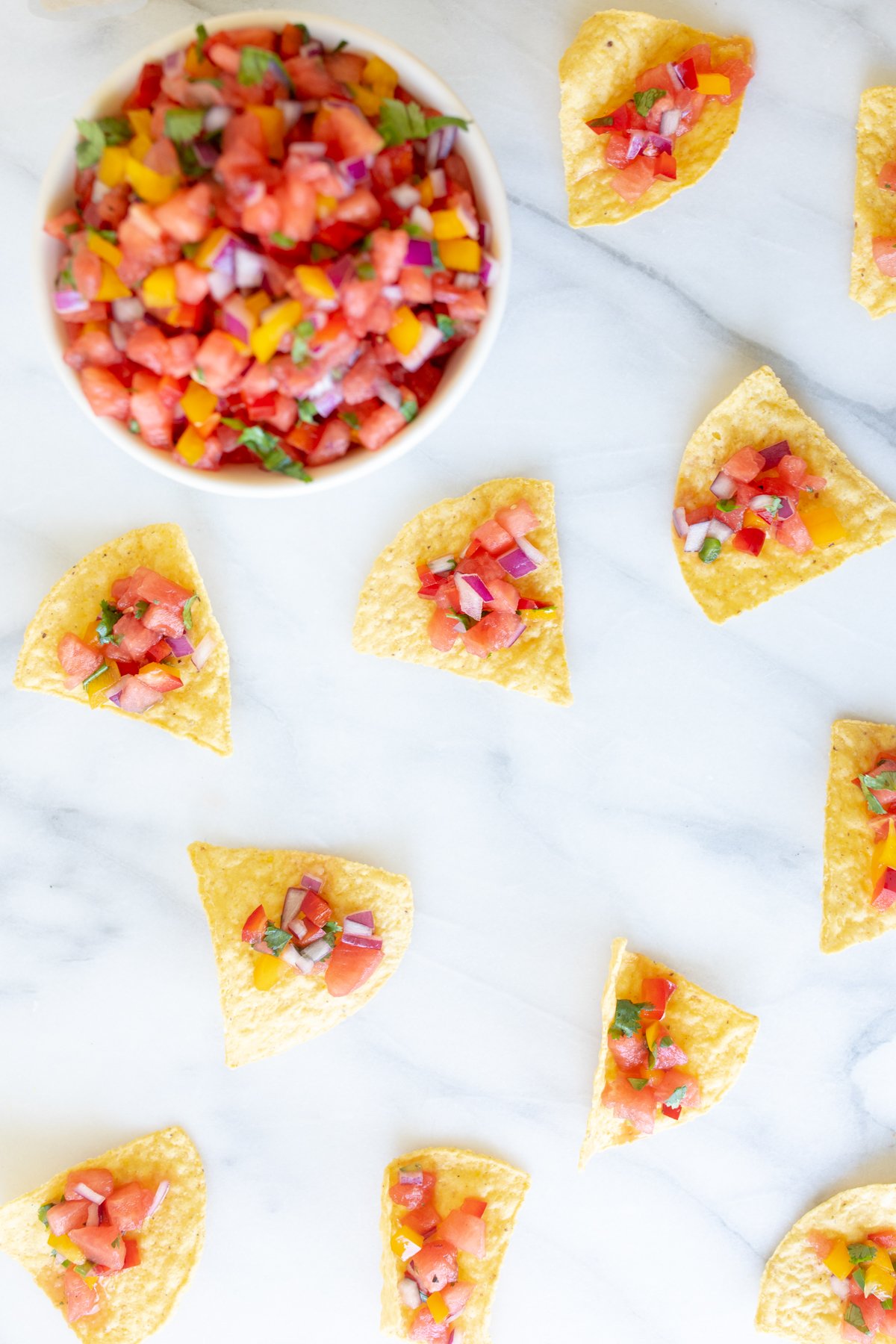 Tortilla chips topped with fresh salsa recipes arranged neatly around a bowl of salsa on a marble surface.