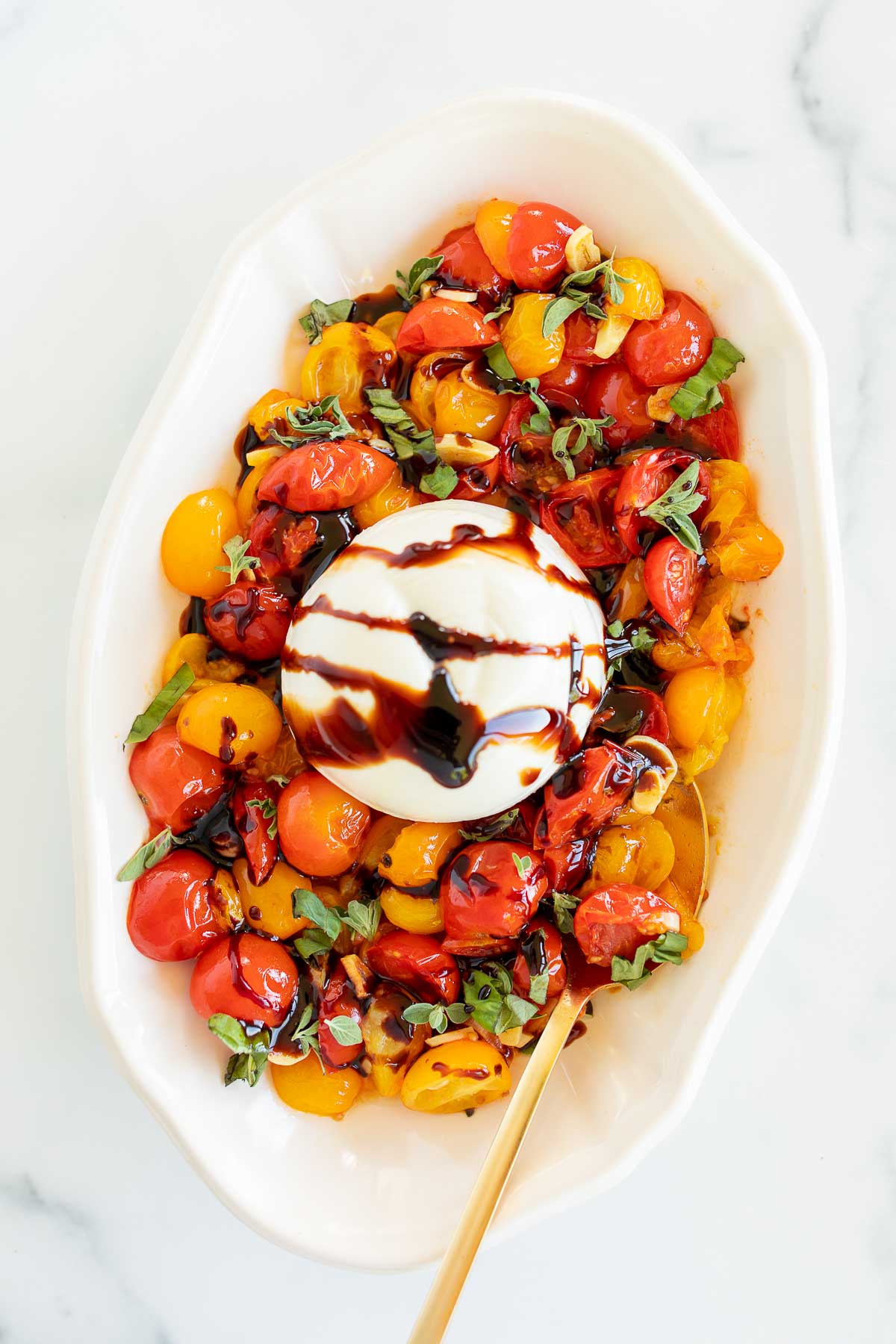 A white dish holds a colorful tomato salad topped with a ball of burrata cheese drizzled with balsamic glaze. Perfect for summer appetizers, the salad is garnished with chopped basil.