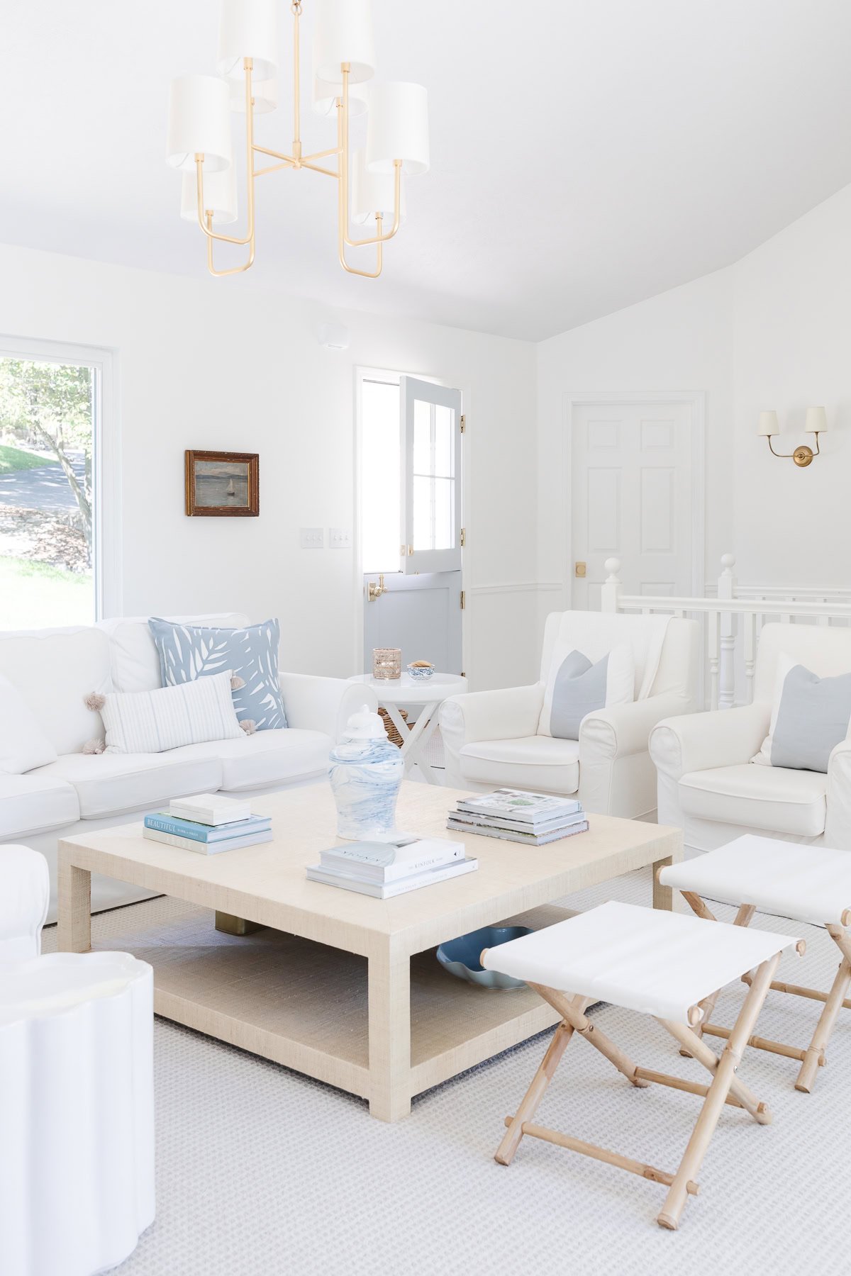 Bright living room with white furniture, a beige coffee table, and decorative throw pillow covers. A pendant light hangs above a small wooden stool. Books and a sculpture adorn the table, while the door and window provide a scenic backdrop.