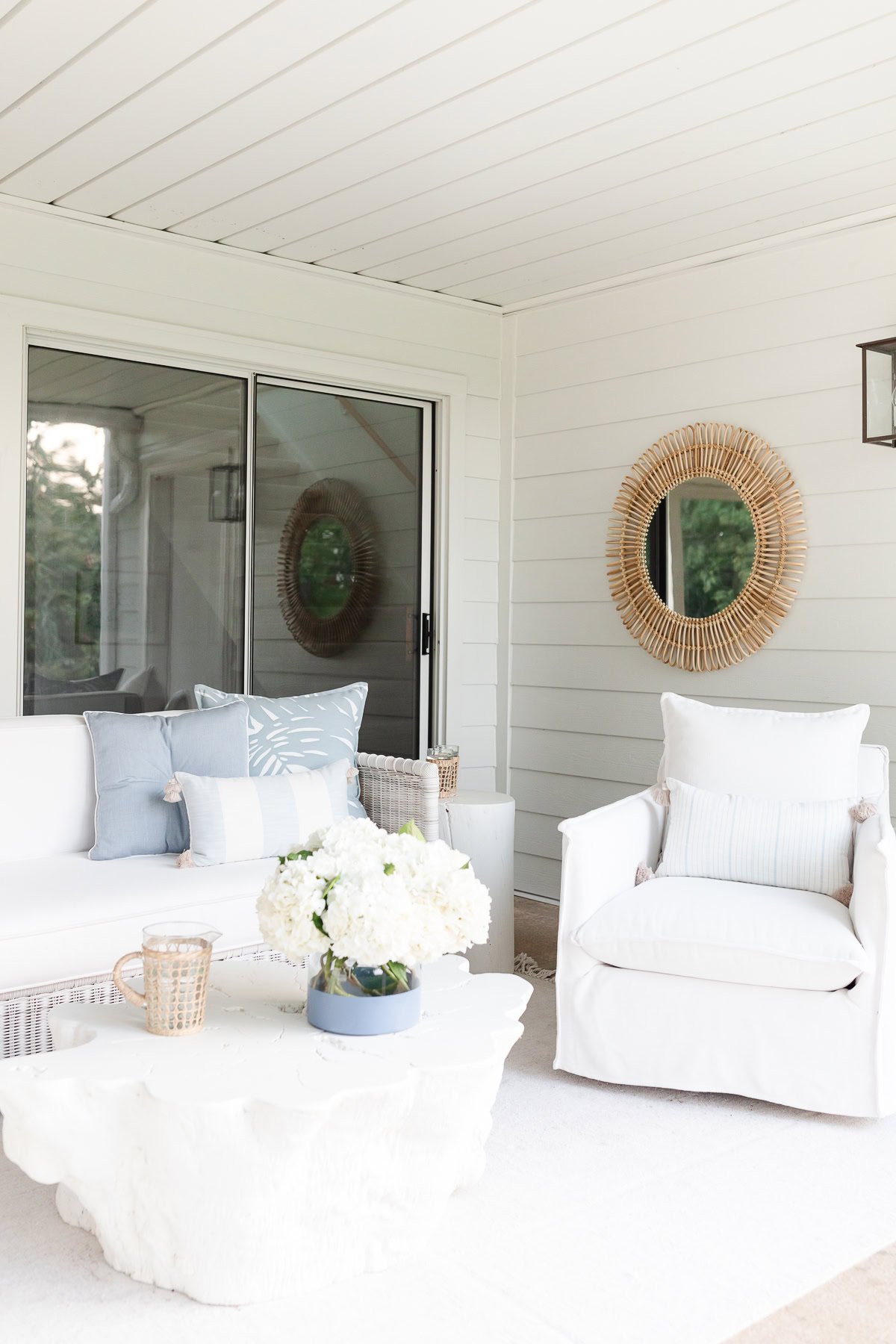 A white outdoor seating area features a sofa and chair adorned with stylish throw pillow covers, a stone coffee table with flowers, a circular mirror on the wall, and elegant glass sliding doors.