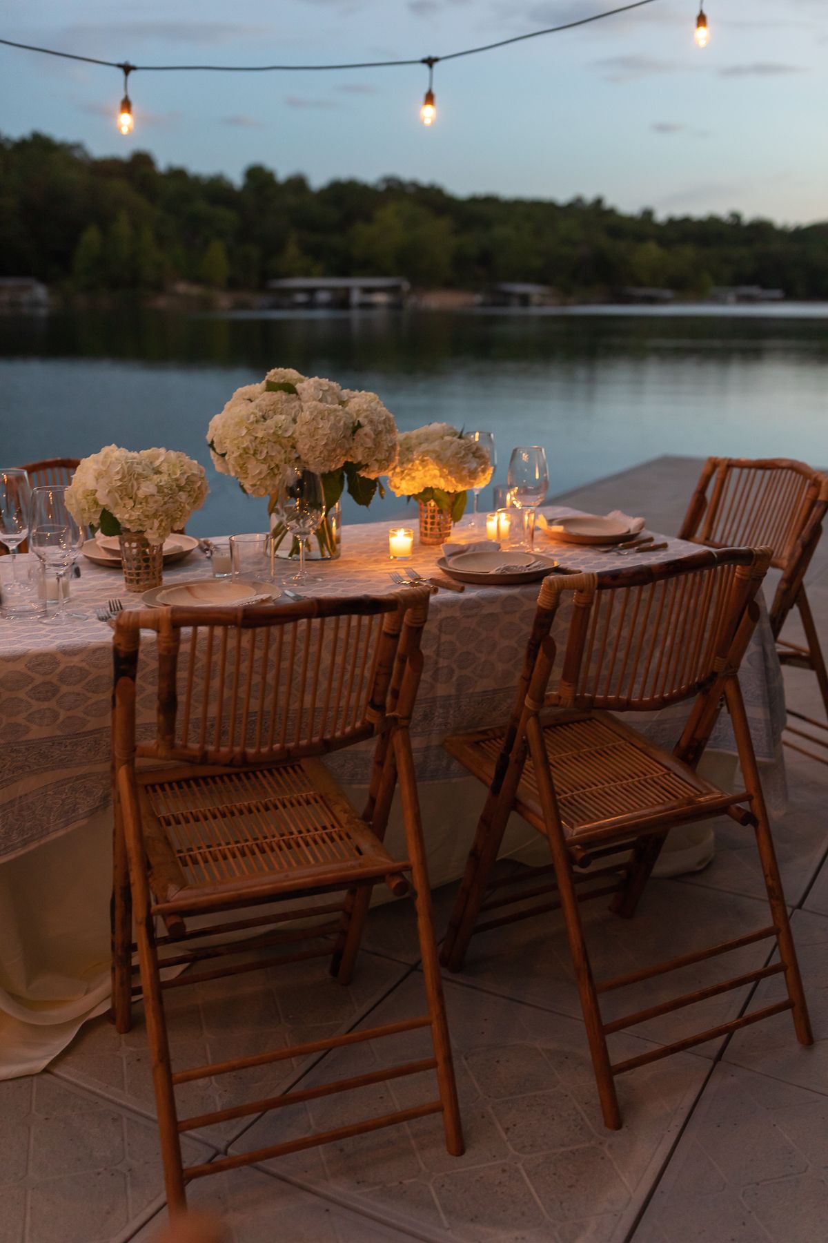 outdoor string lights set up over a dining table on a dock, water in the background