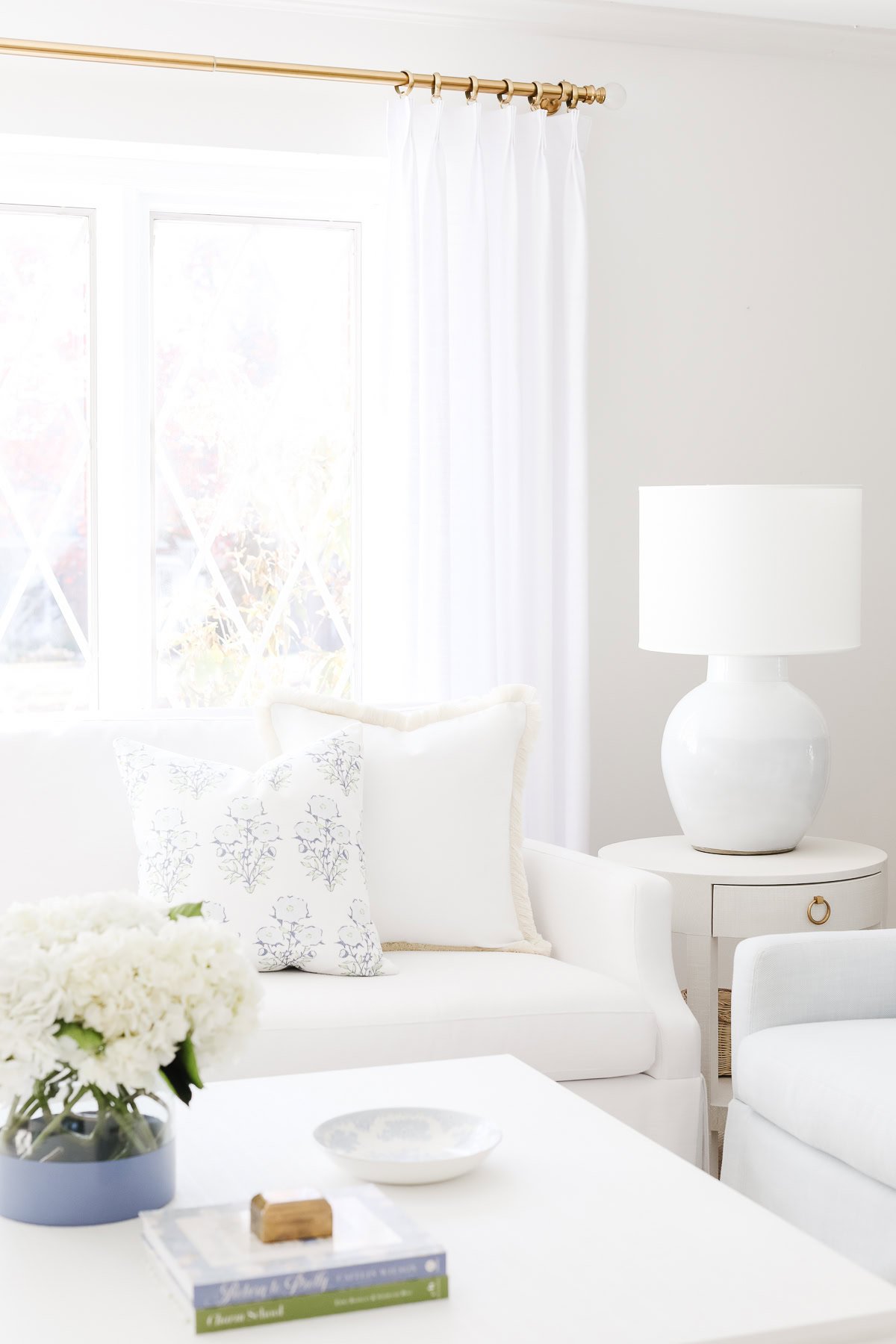 Bright living room with white furniture, a side table with a white lamp, and floral throw pillow covers accenting the cushions. A vase of white flowers graces the coffee table. Large windows provide natural light.