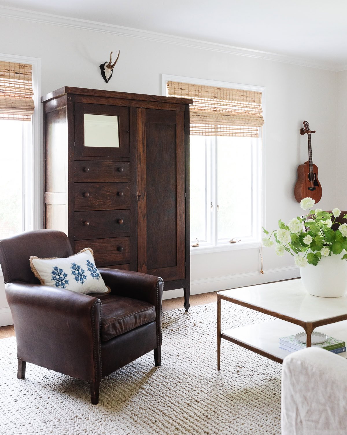 Sala de estar elegante con un sillón de cuero adornado con elegantes cubiertas de almohadas, un gabinete de madera oscura, guitarra montada en la pared y mesa de café. Grandes ventanas con persianas de bambú iluminan el espacio.