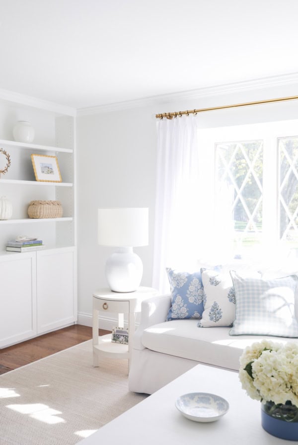 Bright living room with white walls, sofa, and side table. Blue patterned throw pillow covers add a stylish touch alongside the lamp and the window with diamond panes. Shelves with decorative items are neatly arranged on the left.