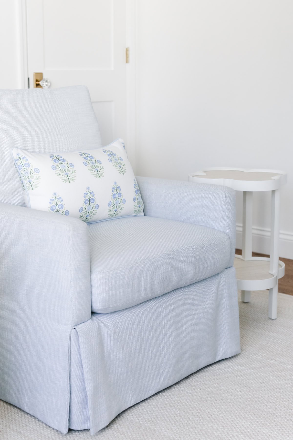 Light blue armchair with floral throw pillow covers next to a small white side table on a carpet in a minimalist room.