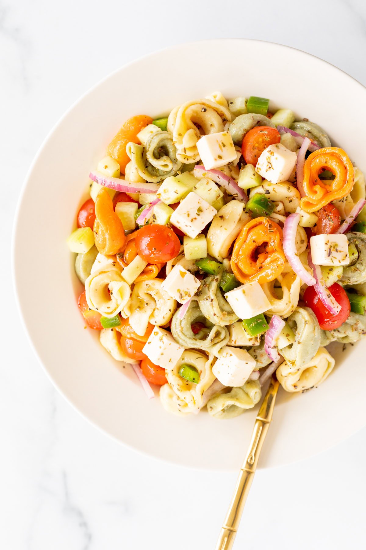 A white bowl filled with pasta salad containing tortellini, cubed cheese, cherry tomatoes, diced cucumbers, red onions, and bell peppers—perfect as one of your pulled pork sides. A gold fork rests on the side.
