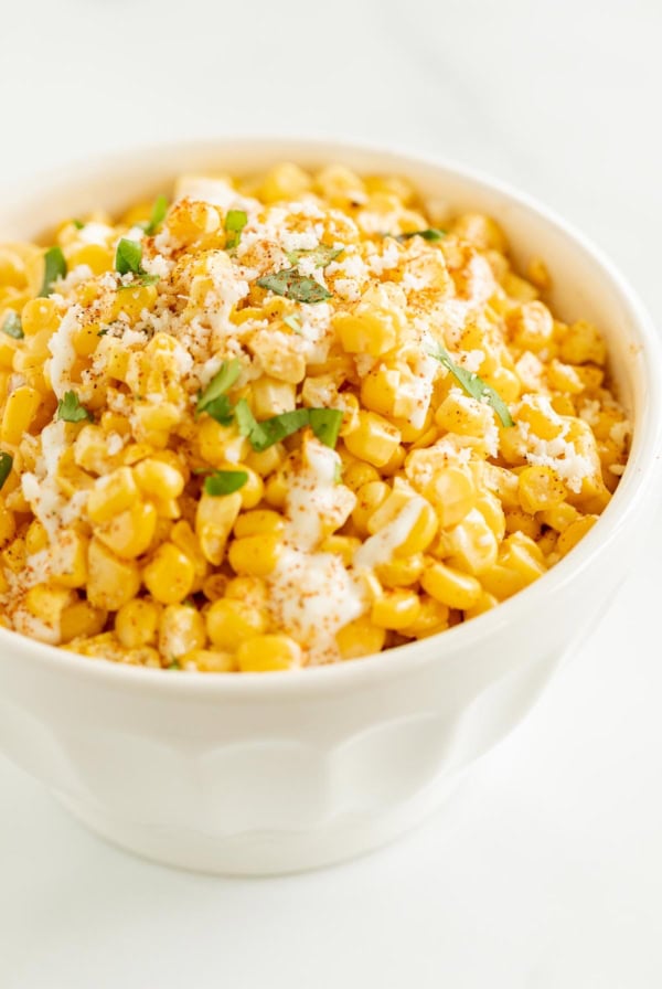 A bowl of seasoned corn topped with herbs and spices, perfect as a side dish for tacos, on a white background.