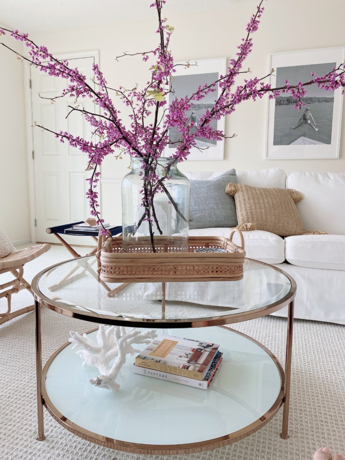 A glass coffee table with nautical inspired coffee table decor and a vase of tall fuchsia blooming branches. 