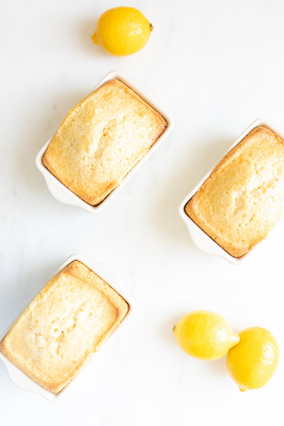 Three baked loaves of lemon bread in white rectangular dishes are arranged on a white surface. Two whole lemons are placed beside the top and bottom right loaves, making for a delightful display of lemon desserts.