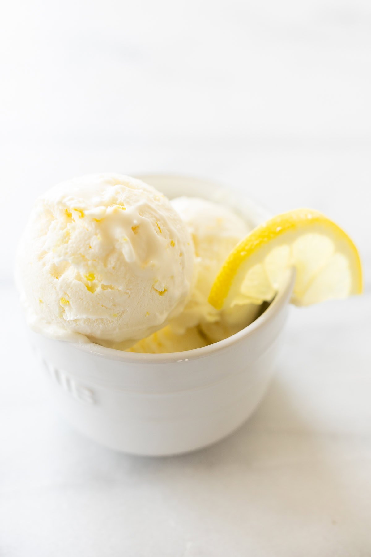 A white ceramic bowl holds two scoops of lemon ice cream, a perfect example of delightful lemon desserts, garnished with a slice of lemon on the edge.