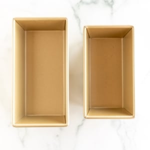 Two different gold loaf pan sizes on a white marble surface